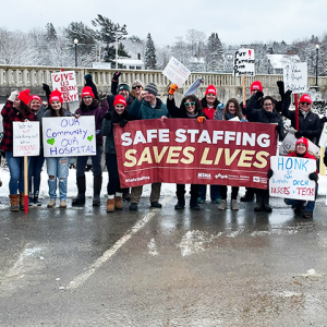 Group photo of RNs and techs by the rally line along the Bad Falls bridge, Machias. Many signs, large banner "Safe staffing saves lives"