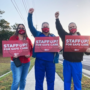 Three nurses outside with raised fists hold signs "Staff Up for Safe Care"