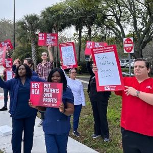 Large group of nurses in blue scrubs outside hospital holding signs "HCA Put Patients Over Profits"
