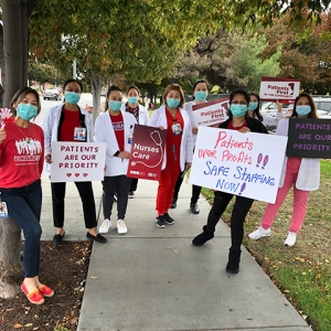 Large group of nurses outside hospital hold signs "Patients First"