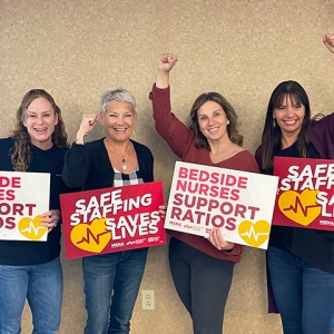Four nurses with raisded fists hold signs in support of safe staffing ratios