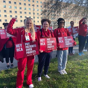 Large group of nurses in blue scrubs outside hospital with raised fists holding signs "HCA Put Patients Over Profits"