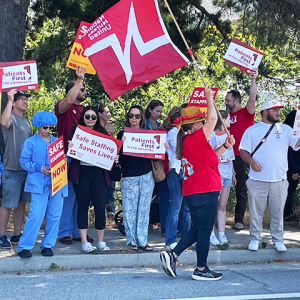 Nurses and community holding signs: "Safe staffing now" "Patients First in the hospital" "Patients First in the hospital" "Safe Staffing Saves Lives"