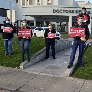 Nurses outside hospital with signs: "Staff up for safe care, end crisis care now"