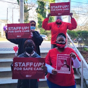 Four nurses outside with raised fists hold signs "Staff Up for Safe Care"