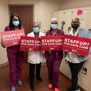 Four nurses inside hospital hold signs "Staff Up for Safe Patient Care"