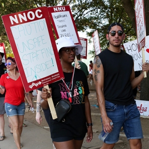 Nurses on picket line, one holds sign "Caution, Unsafe Staffing Inside"