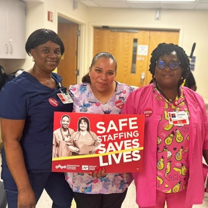 Group of three nurses inside hospital, one hold sign "Saf Staffing Saves Lives"