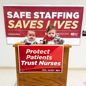 Babies at press event podium above sign "Protect Patients Trust Nurses" with banner in back "Safe staffing saves lives"