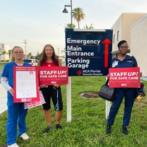 Group of three nurses outside hospital hold signs "People Before Profit" and "Staff Up for Safe Patient Care"