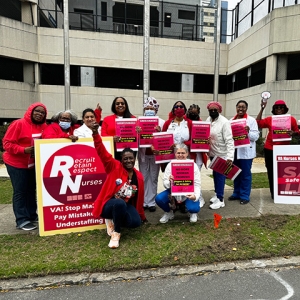 Large group of nurses dressed in red, outside hospital, holding signs "No More Broken Promises: Respect and Safety for All NOW"