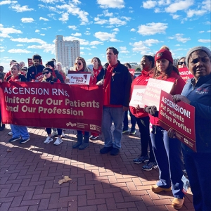 Group of nurses outside holding banner, signs "Ascension, put patients before profits"