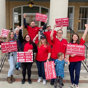 Group of nurses outside in rain clothes holding signs "Some Cuts Don't Heal" and "Fund Care Not Billionaires"
