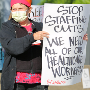 Nurse holds sign "Stop Staffing Cuts! We Need All Health Care Workers"