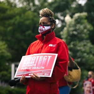 Masked nurse outside holds signs "Protect Nurses, Patients, Public Health"