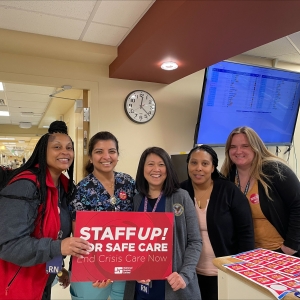 Five nurses inside hospital hold signs "Staff Up for Safe Patient Care"