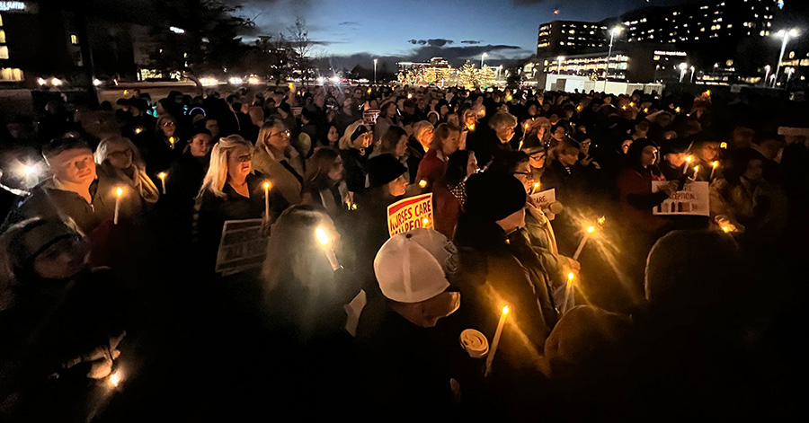 Large group of people outside, at night, holding candles. Some hold signs saying "Nurses Care for All People"