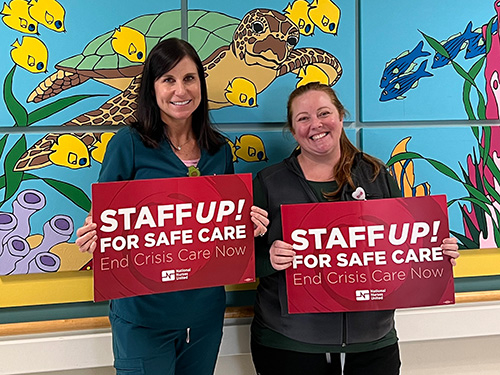 Two nurses smiling, inside hospital, holding signs "Staff Up For Safe Care"