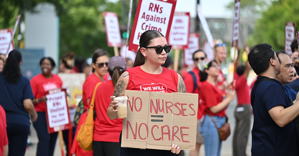 Nurse on picket line holds sign "No Nurses = No Care"