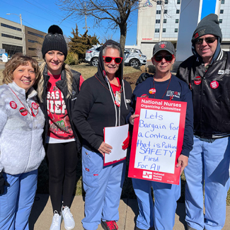 Group of nurses outside hospital, one holding sign "Let's bargain for a contract that puts safety first"