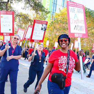 Nurses on picket line, smiling, one holds sign "We Need a Contract NOW!"