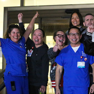 Large group of nurses outside hospital, smiling, with raised fists