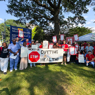 Large group of nurses and community outside hospital, holding signs and banner "MedStar: Stop Cutting Maternal Health"