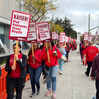 Nurses on picket line outside of hospital, holding signs "Kaiser: Put Patients Over Profits"