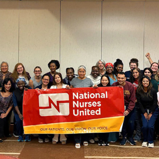 Large group of nurses inside conference room holding banner "National Nurses United"