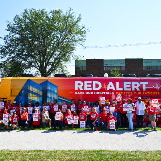 Nurses at Olympa Fields in front of Red Alert Tour bus with signs "Prime RNs United for Our Patients"
