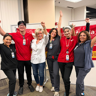 Six nurses inside, side-by-side, smiling, with raised arms/fists