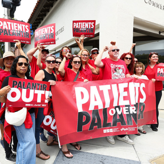 Group of nurses hold signs and banner "Palantir Out" and "Patients Over Palantir"
