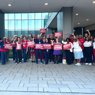 Large group of nurses outside hospital with raised fists, some holding signs "Charity Betrayed"