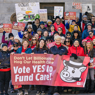 Large group of people holding banner and signs "Don't Let Billionairs Hog Our Health Care: Vote Yes to Fund Care"