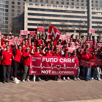 Large group of nurses outside holding banner and signs "Nurses Say: Fund Care Not Billionaires"