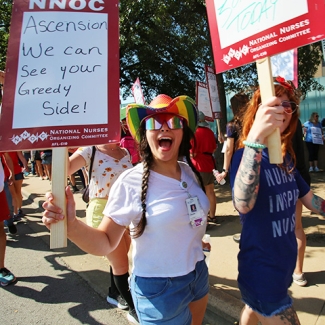 Nurses on picket line, one holds sign "Ascension, we can see your greedy side"