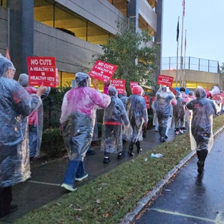 Nurse on picket line in the rain, wearing ponchos, holding signs "No Cuts: A Healthy VA = Health Veterans"