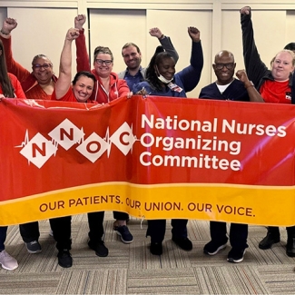 Group of nurses smiling, with raised fists, holding banner "National Nurses Organizing Committee - Our Patiens, Our Union, Our Voice""