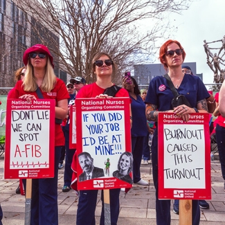 Three nurses stand side-by-side outside holding signs criticizing UMC/LCMC