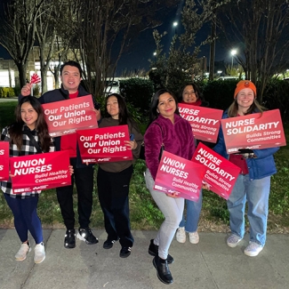 Group of nurses outside hospital at night, holding signs and banner "Our Patients, Our Union, Our Rights"