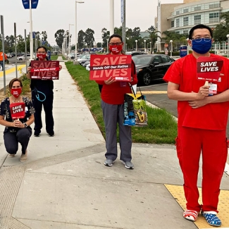 Nurses hold signs "Save Lives" and variations "Hands Off Our Ratios" "Safe Staffing Now" "PPE Now"