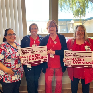 Group of four nurses inside hospital hold signs "Save Our Community Hospital: Save Hazel Hawkins"