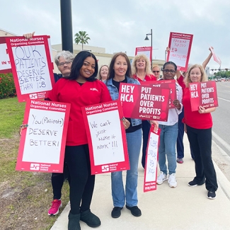 Large group of nurses in blue scrubs outside hospital holding signs "HCA Put Patients Over Profits" and calling for improved working conditions