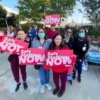 Group of nurses outside, smiling, holding signs "Safe Staffing Now"