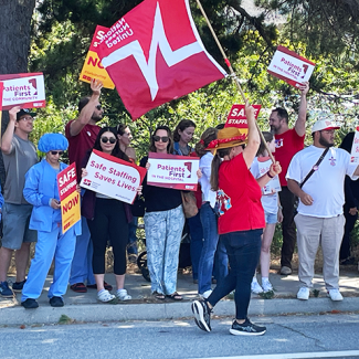Nurses and community holding signs: "Safe staffing now" "Patients First in the hospital" "Patients First in the hospital" "Safe Staffing Saves Lives"