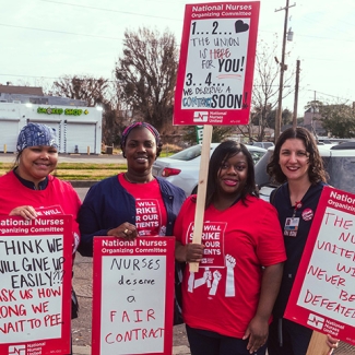 Four nurses standing outside smiling, holding signs calling for a fair contract