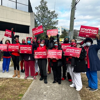 Large group of nurses outside hospital hold signs "Staff Up for Safe Patient Care"