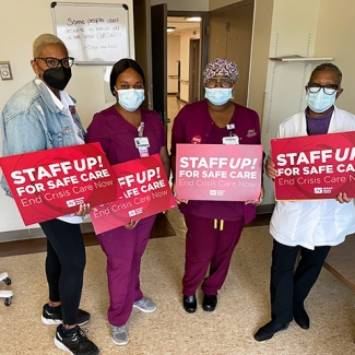 Four nurses inside hospital hold signs "Staff Up for Safe Patient Care"