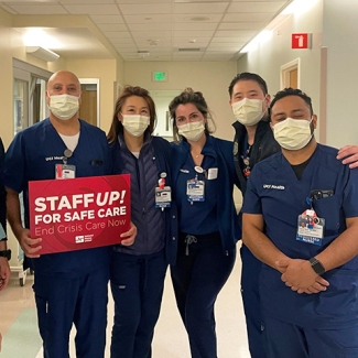 Goup of nurses inside hospital, one holds sign "Staff Up for Safe Care"