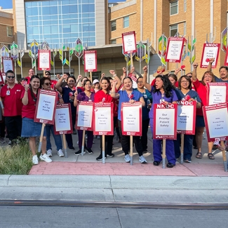 Large group of nurses outside hospital with fists raised, signs that read "Patients are our top priority"